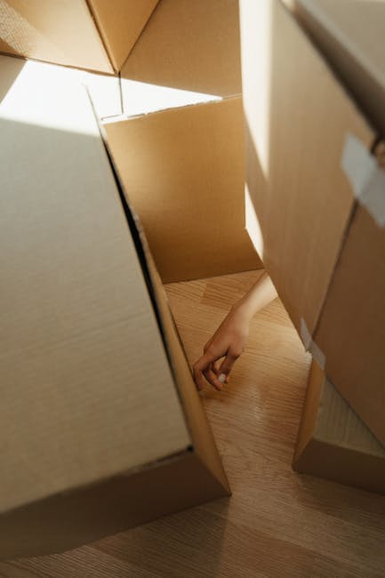 View from inside a filled moving van or storage area showing open cardboard boxes stacked on top of each other, some with visible packing materials like paper or plastic wrapping. A person's hand is r