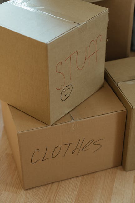 View from inside a filled moving van or storage area showing open cardboard boxes stacked on top of each other, some with visible packing materials like paper or plastic wrapping. A person's hand is r