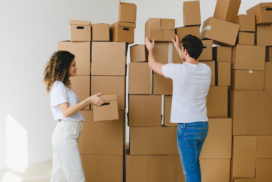Two removal professionals from Removal Van Bayswater are inside a room engaged in home relocation activities. The woman, on the left, has curly hair and is holding a small cardboard box, preparing to stack or move it. The man, on the right, is wearing a white t-shirt and blue jeans and is actively lifting or arranging a larger cardboard box on top of a pile. The background consists of numerous neatly stacked cardboard boxes of varying sizes, some sealed with packing tape, indicating a packing and moving process. The boxes are placed against a plain white wall, with natural light illuminating the scene, suggesting a daytime setting. There is a focus on the careful handling of packing materials and the organization involved in furniture transport and packing during a house move, with equipment such as the boxes and possibly a trolley nearby, in line with professional removal services.